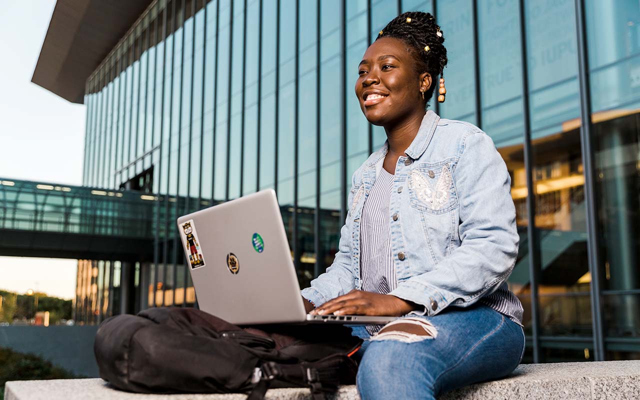 A female student sits outside the IU Indianapolis Campus Center and smiles while using her laptop.